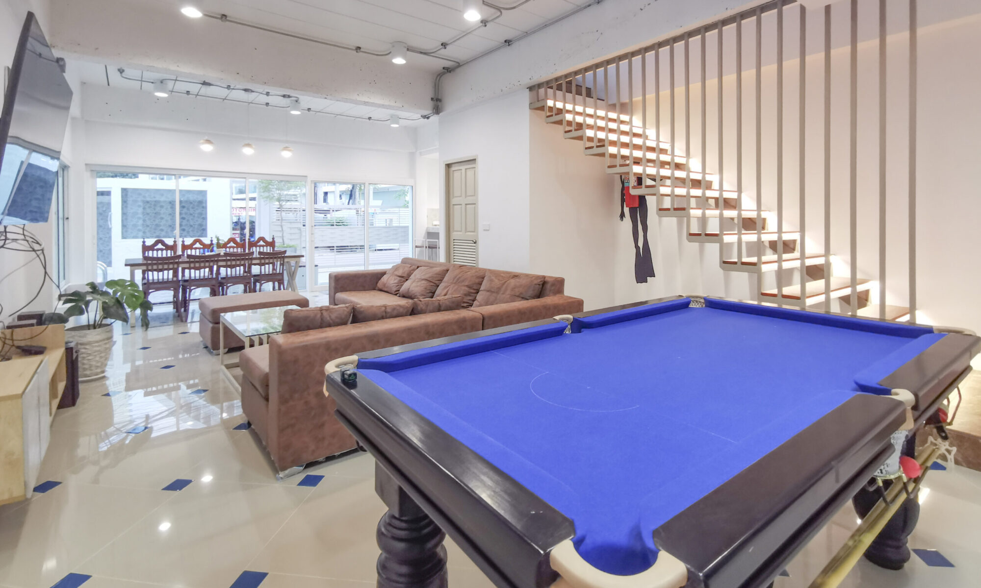 Contemporary recreation room at Thepprasit Cosy Twin blue-felt pool table, leather seating, wall-mounted TV, and illuminated staircase with wooden slats.